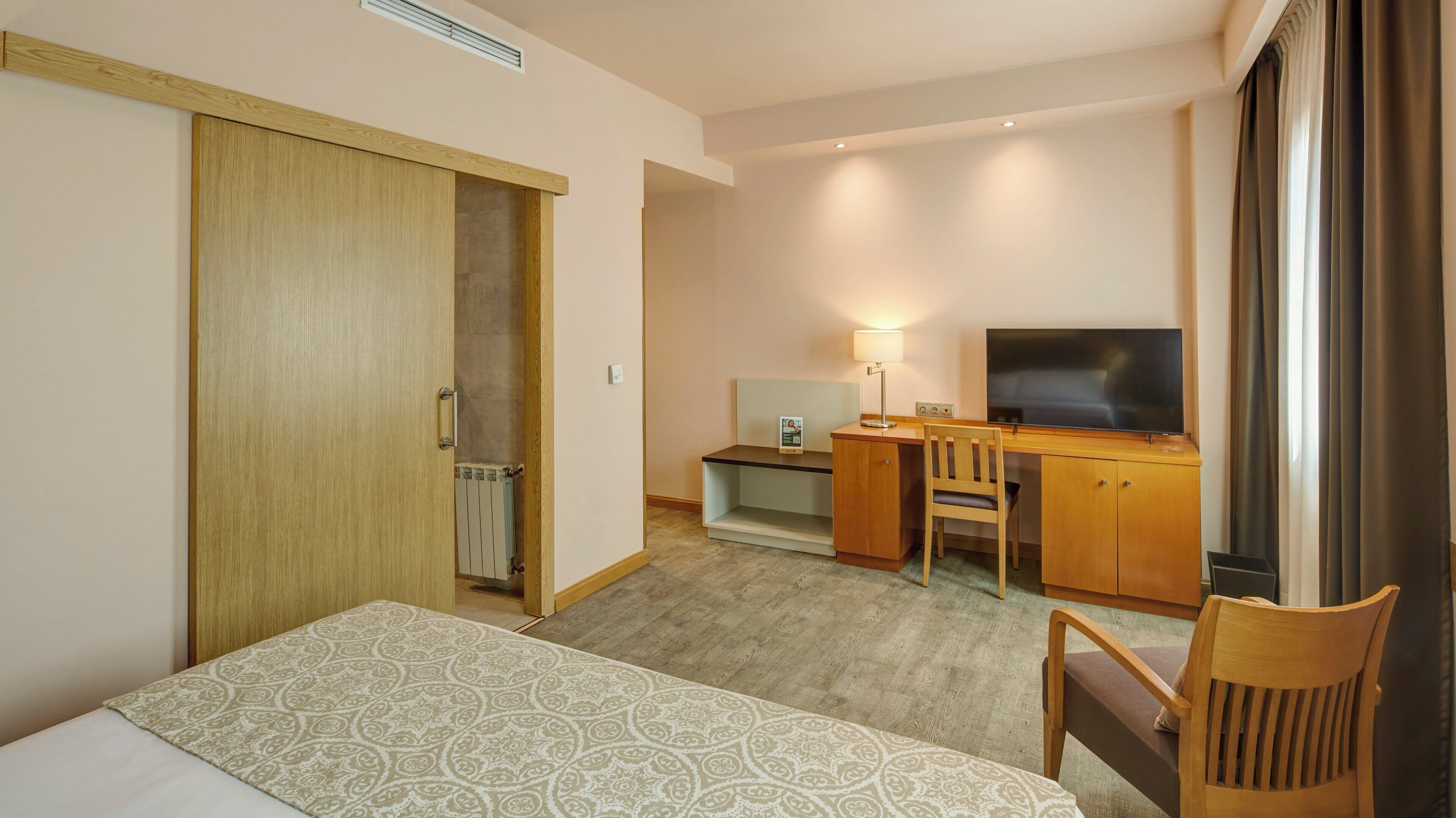 A hotel room with a bed, a desk with a chair, a television, and a wardrobe.