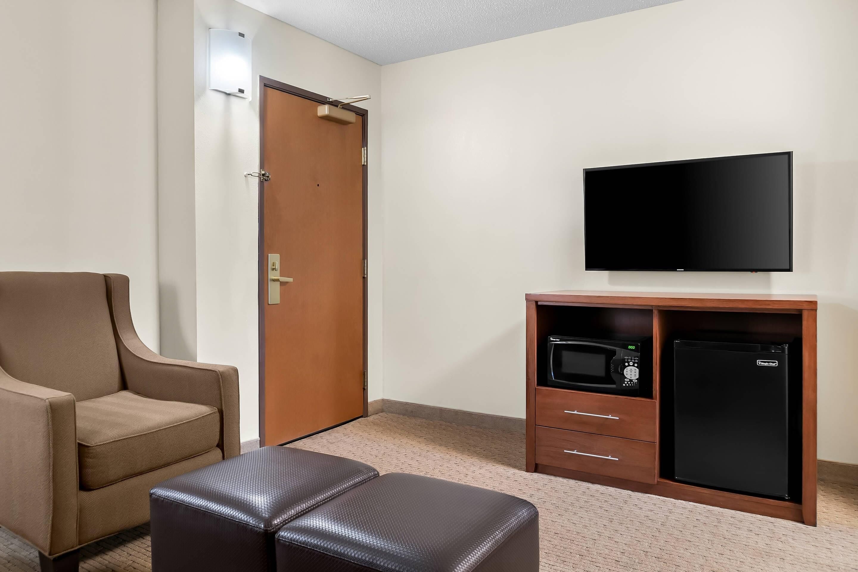 Habitación de hotel con televisión de pantalla plana, minibar, un sillón marrón y un pequeño otomaní.