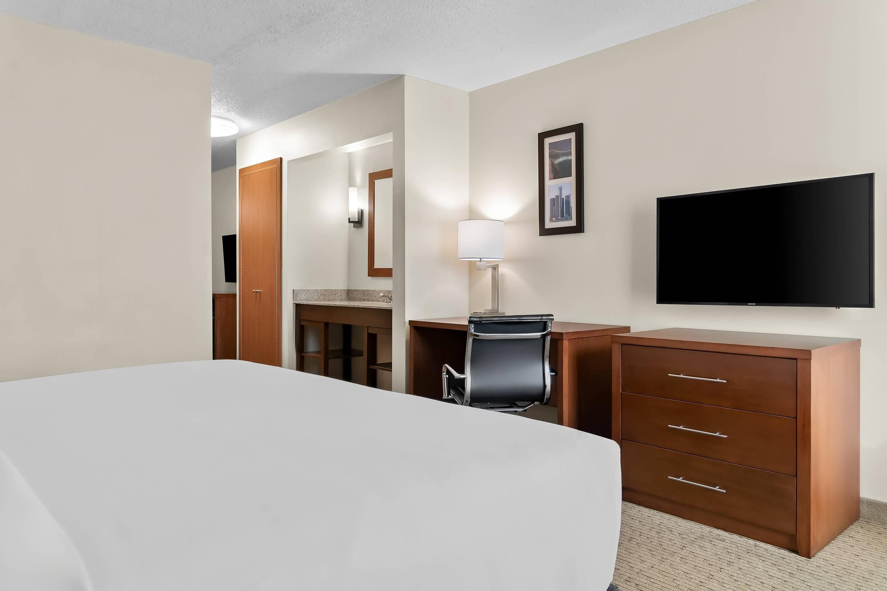 A hotel room with a bed, desk, chair, dresser, TV, and a mirror.