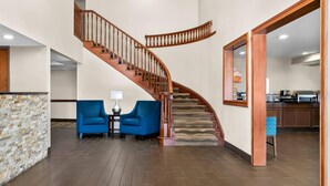 A modern interior with a curved wooden staircase, two blue armchairs, a small table with a lamp, and a kitchen area in the background. - Comfort Inn & Suites Taylor - Detroit (Taylor)