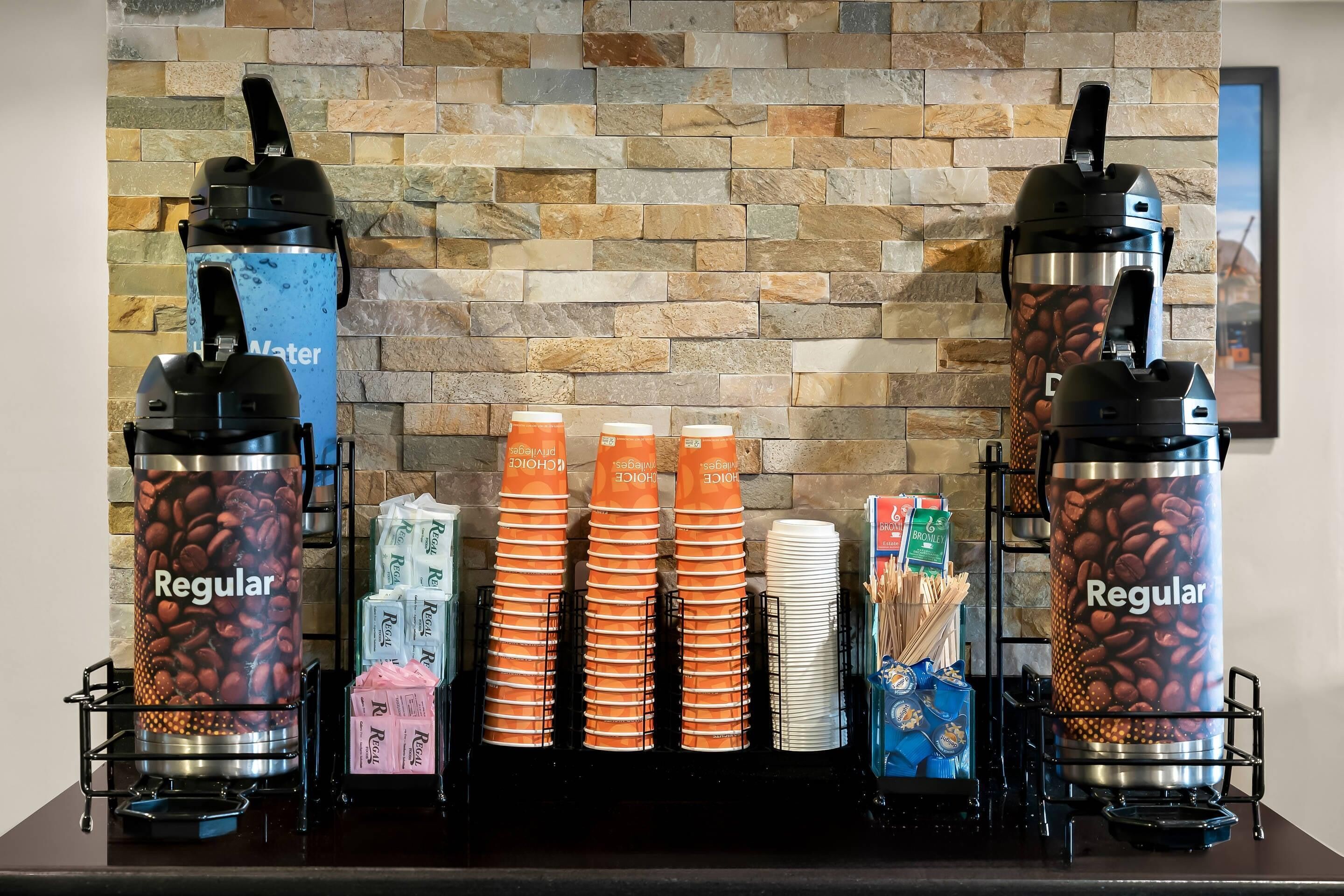 Un espace dédié au café, équipé de deux distributeurs de café, de piles de gobelets orange et blancs, et d’une variété de dosettes de café.