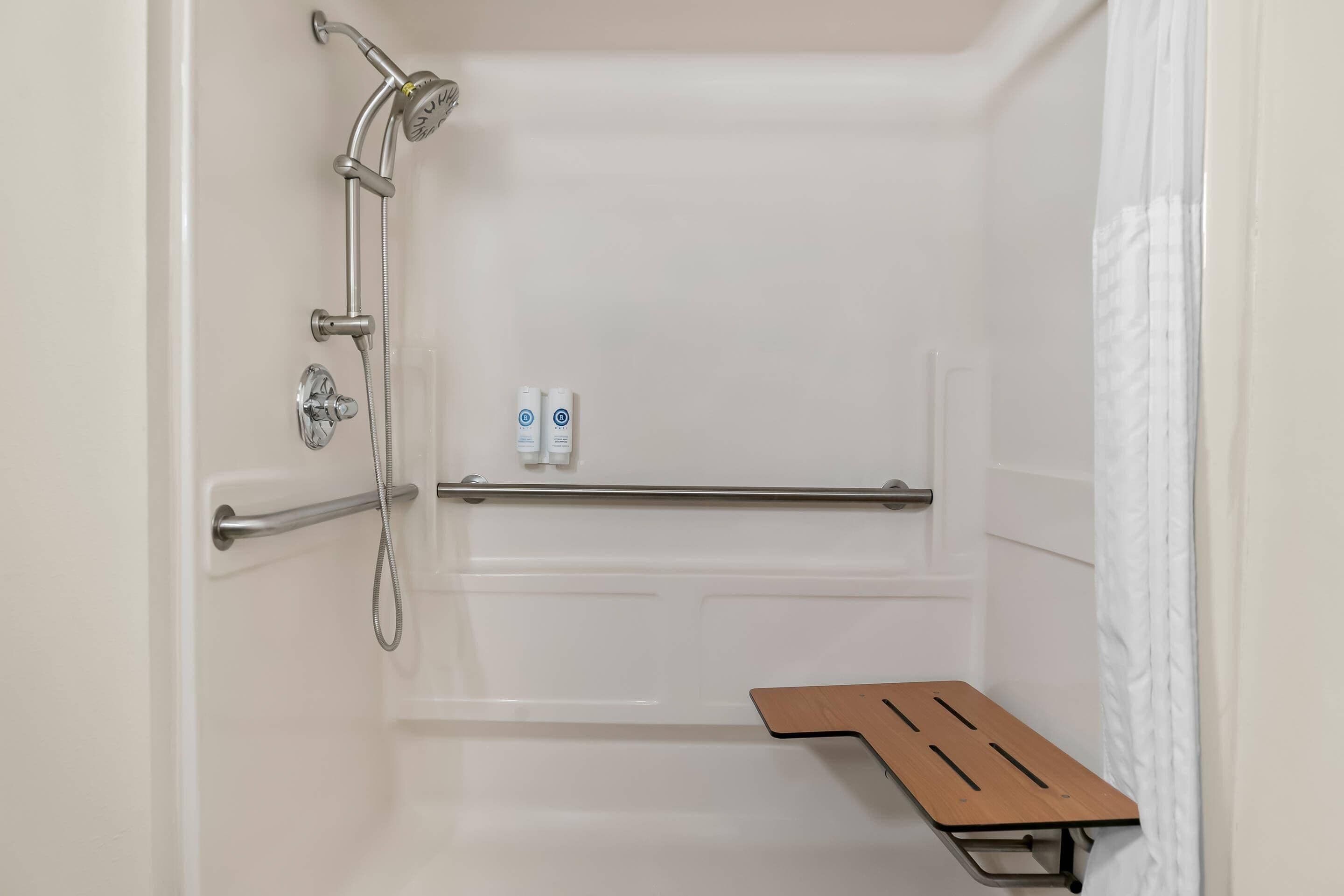 A shower area with a handheld showerhead, a handheld shower attachment, a shower control panel, and a wooden bench.