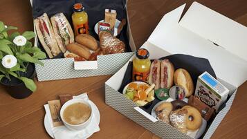 Zwei offene Kisten mit Frühstücksartikeln, darunter Sandwiches, Gebäck, Früchte und Getränke, auf einem Holztisch.