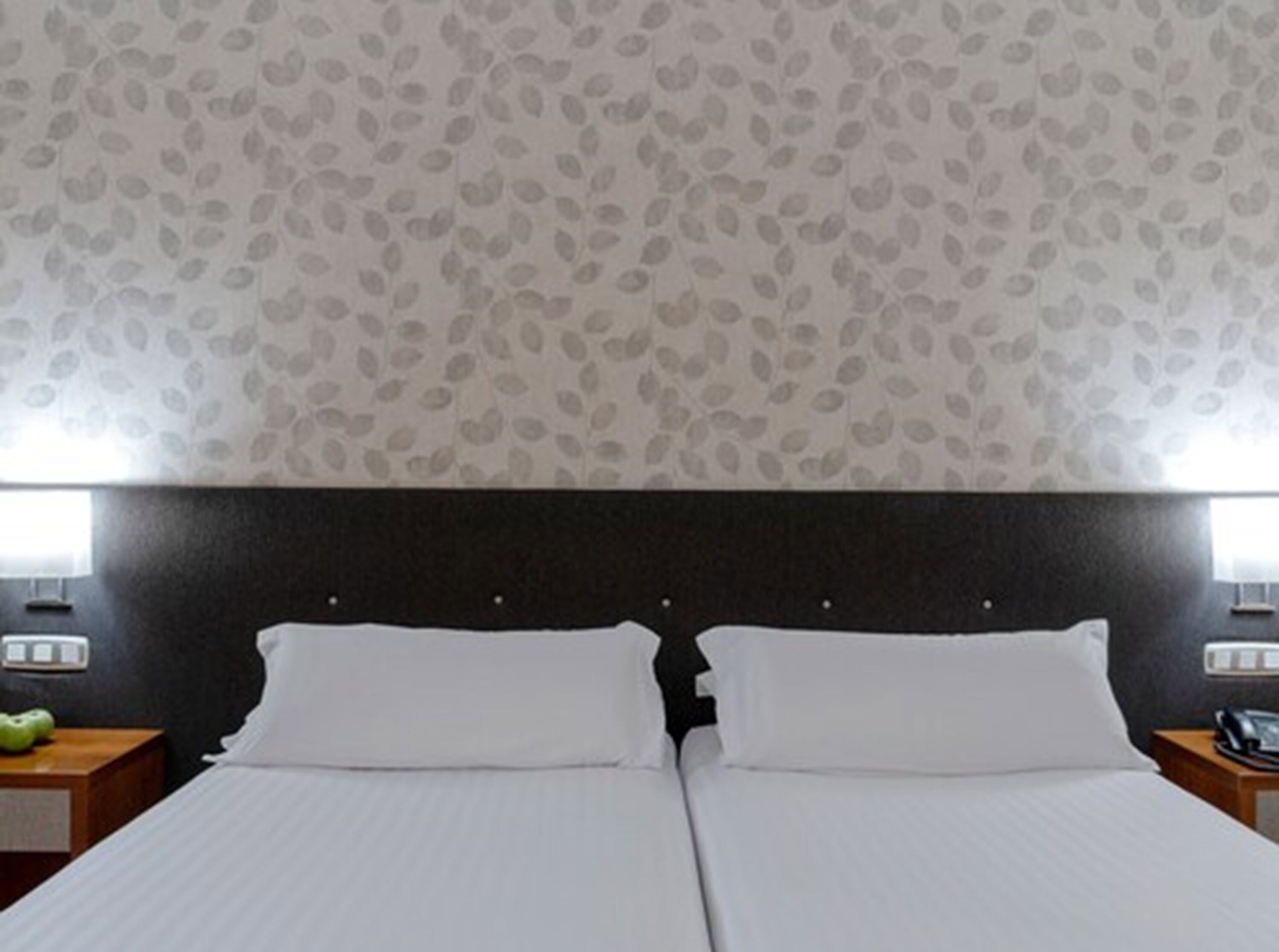 A double bed with white linens, a dark headboard, and a leaf-patterned wallpaper.