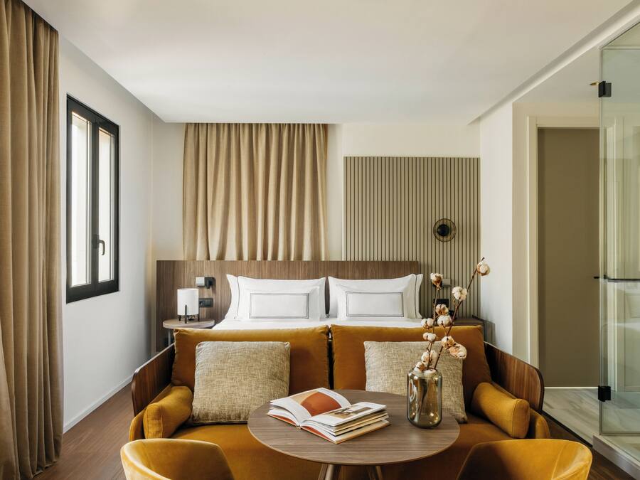 A hotel room with a bed, a sofa, a small table with a book and vase, and a glass shower enclosure.