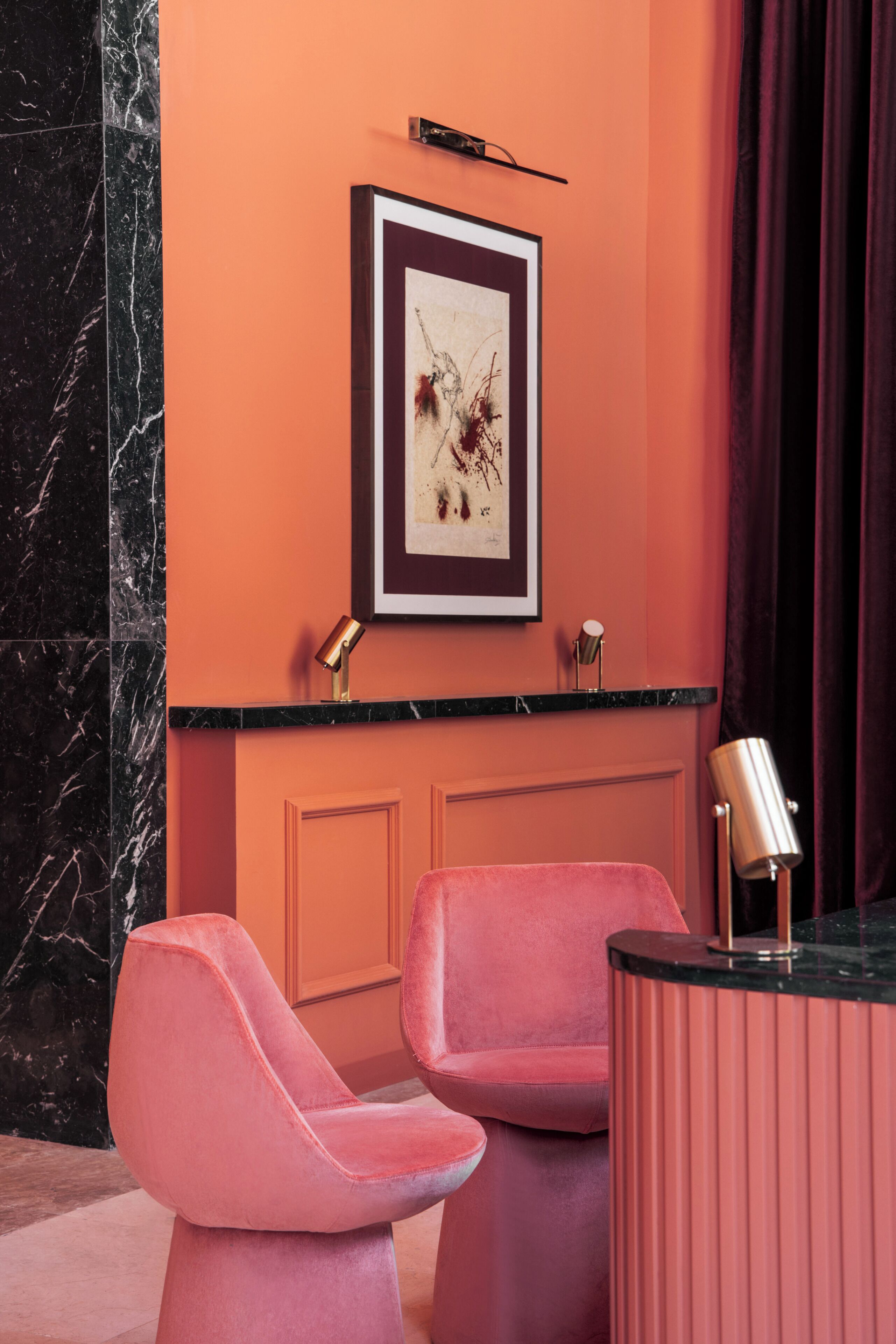 Una habitación con dos sillas rosadas, una pared de mármol, una pared de acento naranja, una obra de arte enmarcada y una encimera negra con una lámpara dorada.