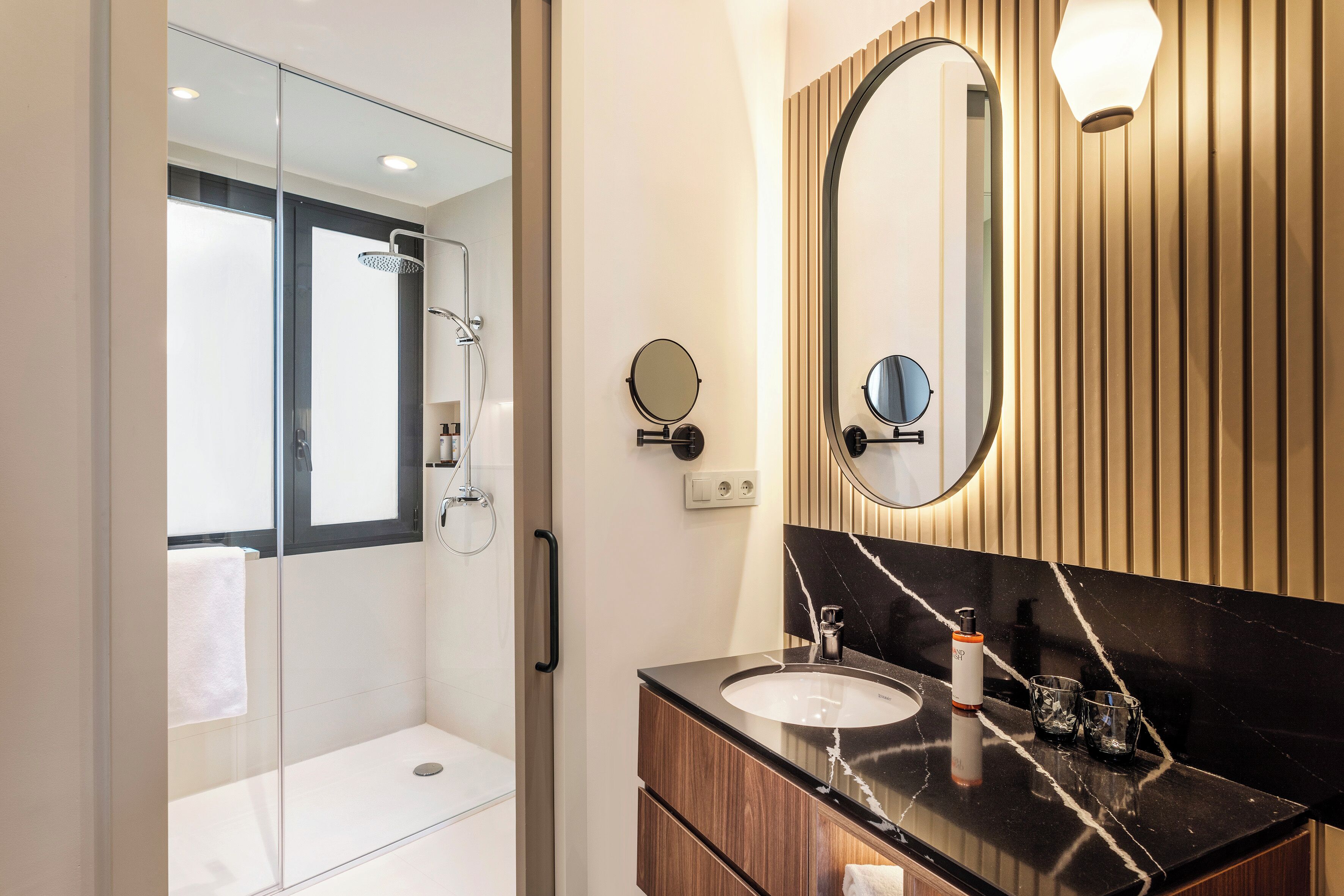 Un baño moderno con cabina de ducha de vidrio, tocador de madera con encimera de mármol negro y un espejo grande con un fondo de panel de madera.