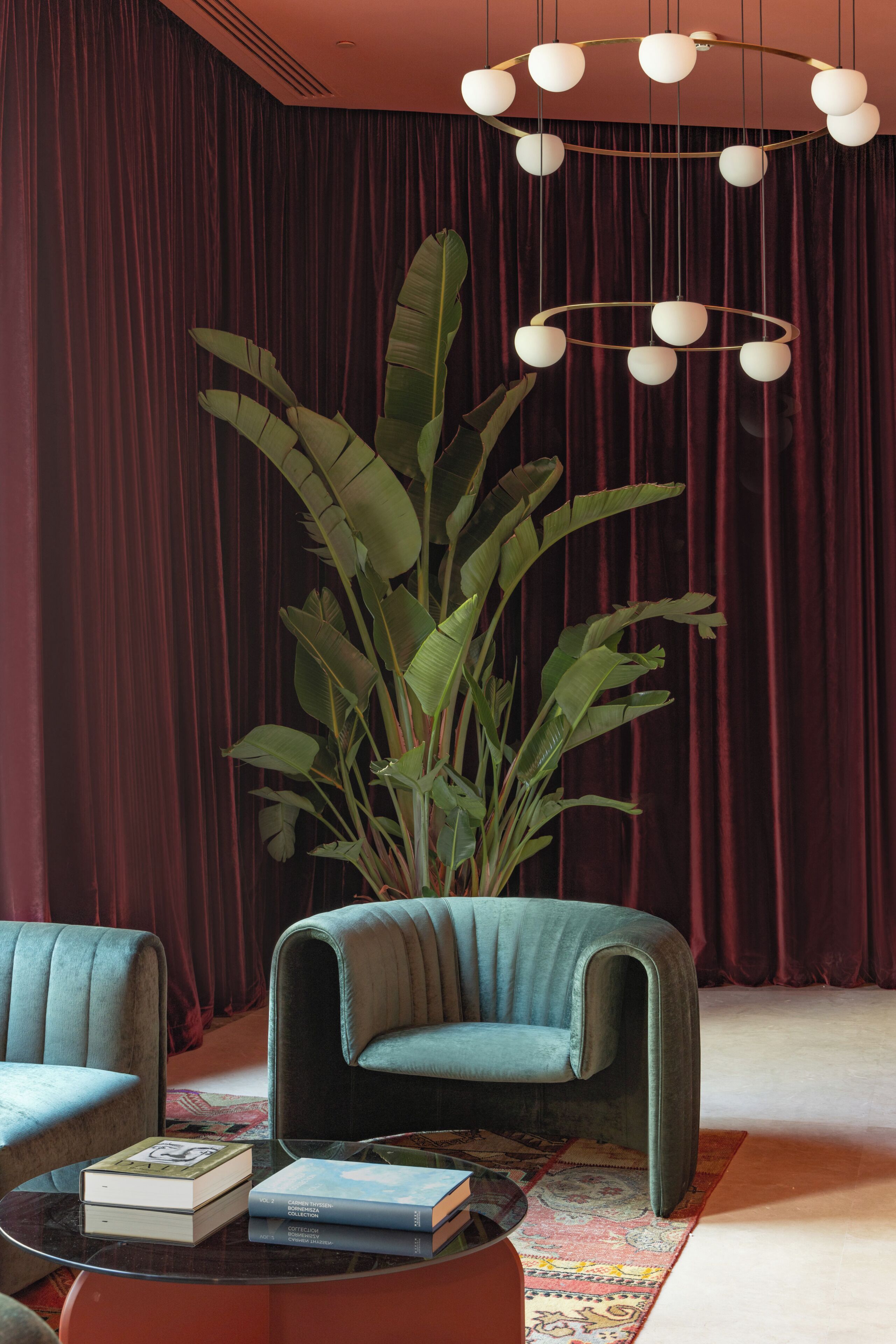 A room with a large plant, a blue armchair, a round coffee table with books, and a red curtain backdrop.