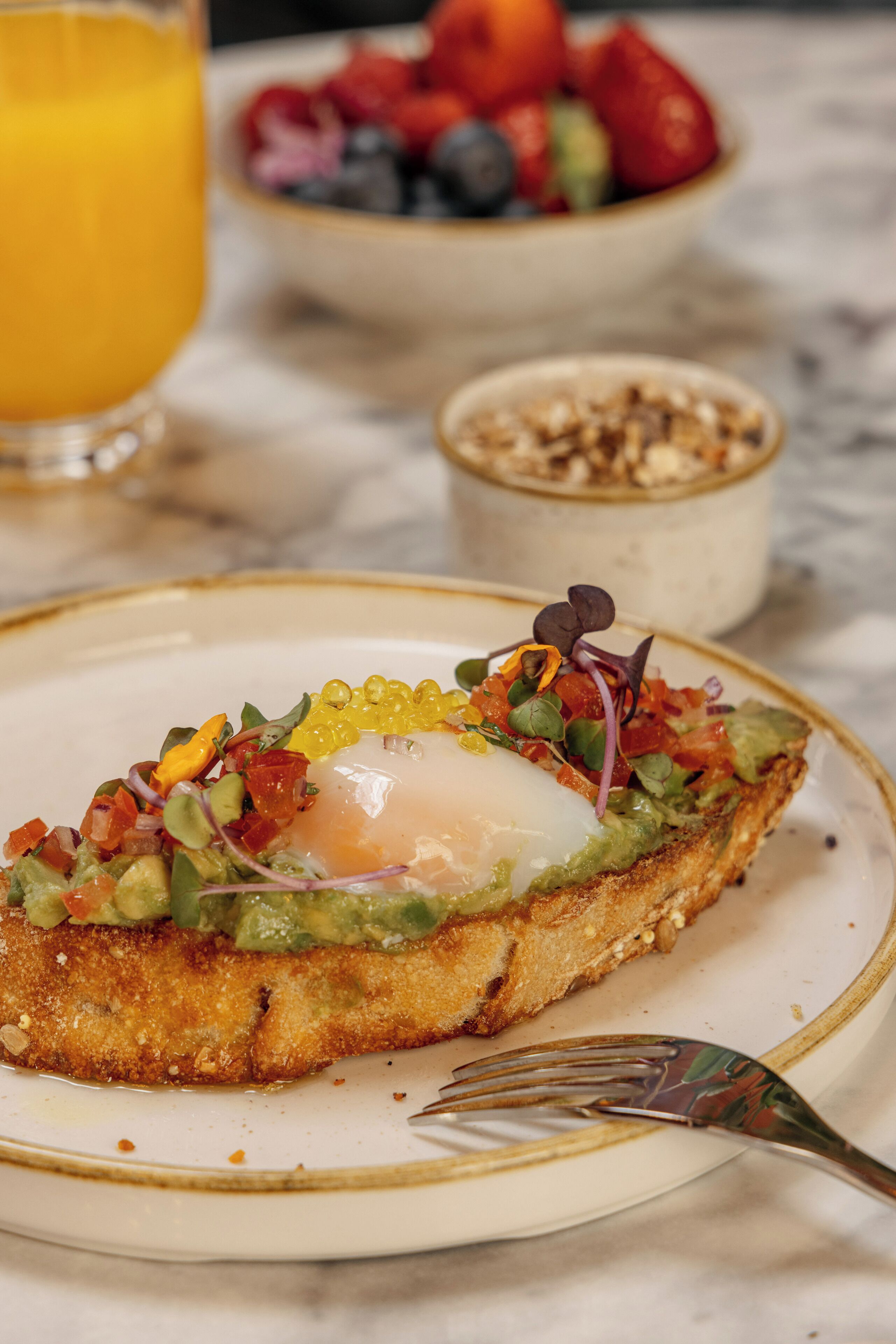 Un plato de desayuno con un huevo pochado, aguacate y verduras de hoja, un tazón de bayas mixtas y un vaso de jugo de naranja.