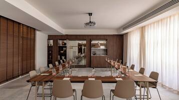 Une salle de conférence équipée d’une longue table, de chaises, d’un projecteur et offrant une vue sur un espace cuisine.