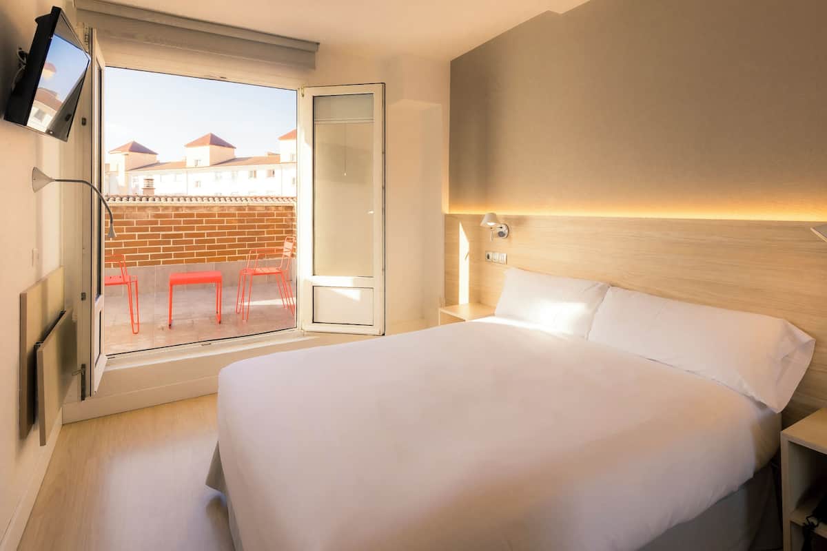 A hotel room with a bed, a TV mounted on the wall, a window with a view of buildings, and a red chair outside.