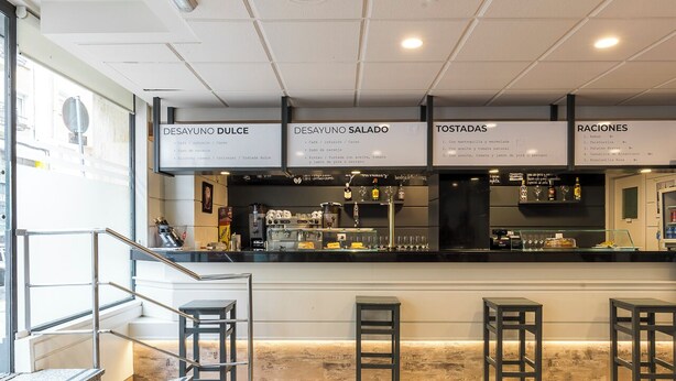 Un bar moderno con barra, taburetes y un panel de menú que muestra opciones como 'Desayuno Dulce' y 'Desayuno Salado'.