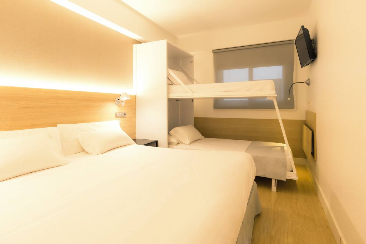 A hotel room with two beds, a bunk bed, a TV mounted on the wall, and a window providing natural light.