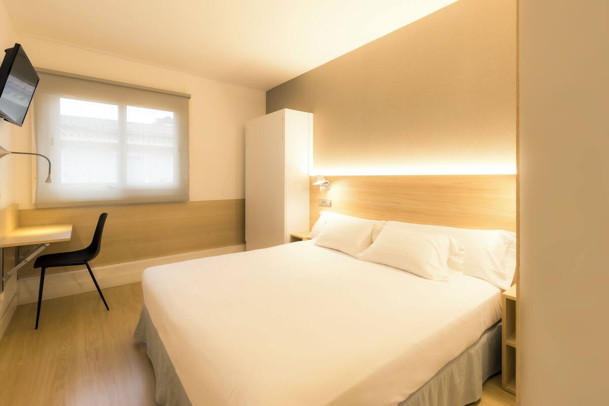 A hotel room with a bed, a desk, a chair, a TV, and a window.