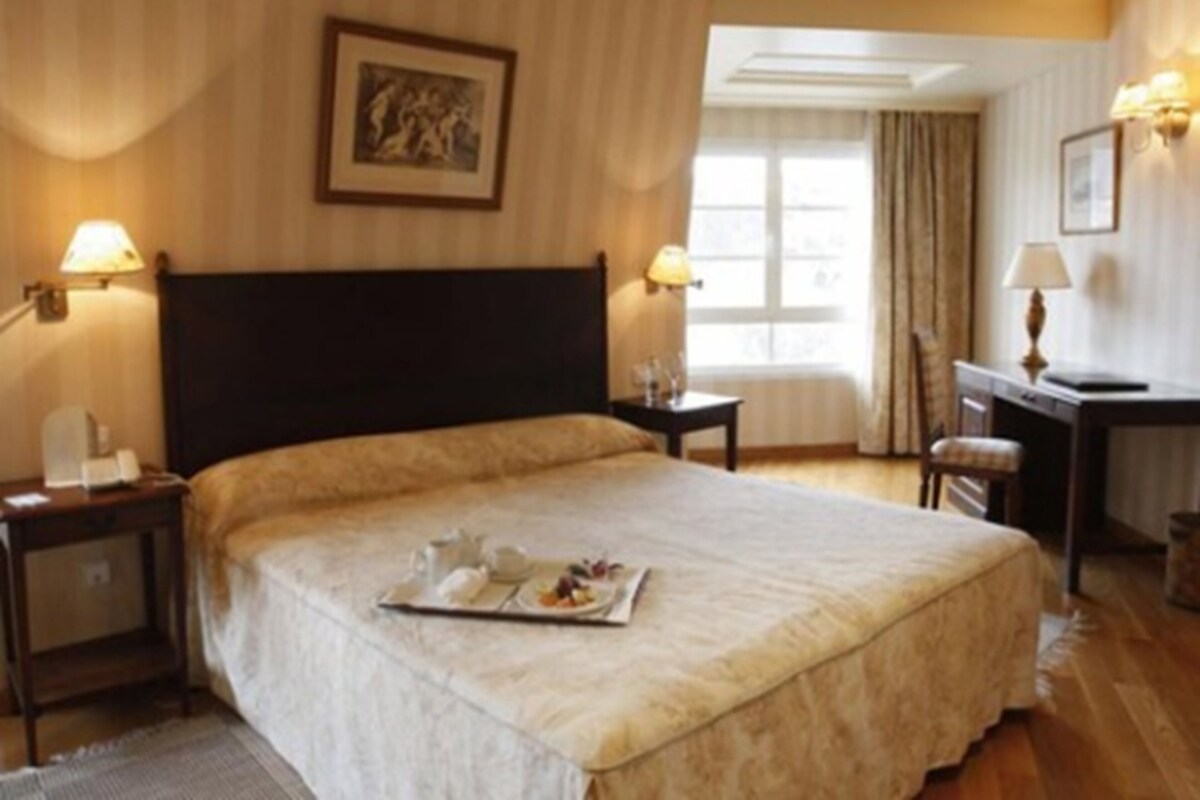 Quarto de hotel com uma cama grande, mesas de cabeceira, uma escrivaninha e uma janela com cortinas.