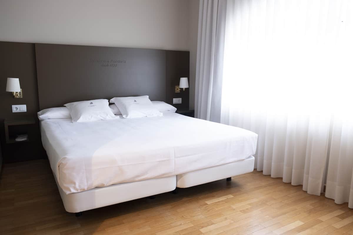 Un lit bien fait dans une chambre d’hôtel, avec un plancher en bois, deux tables de chevet avec des lampes et une grande fenêtre avec des rideaux.