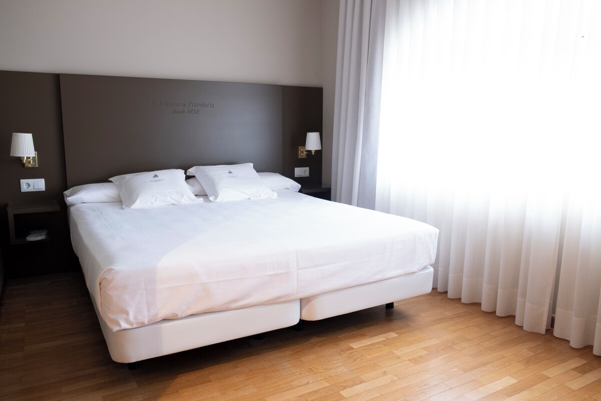 Cama arrumada em quarto de hotel, com piso de madeira, duas mesas de cabeceira com abajures e uma janela grande com cortinas.