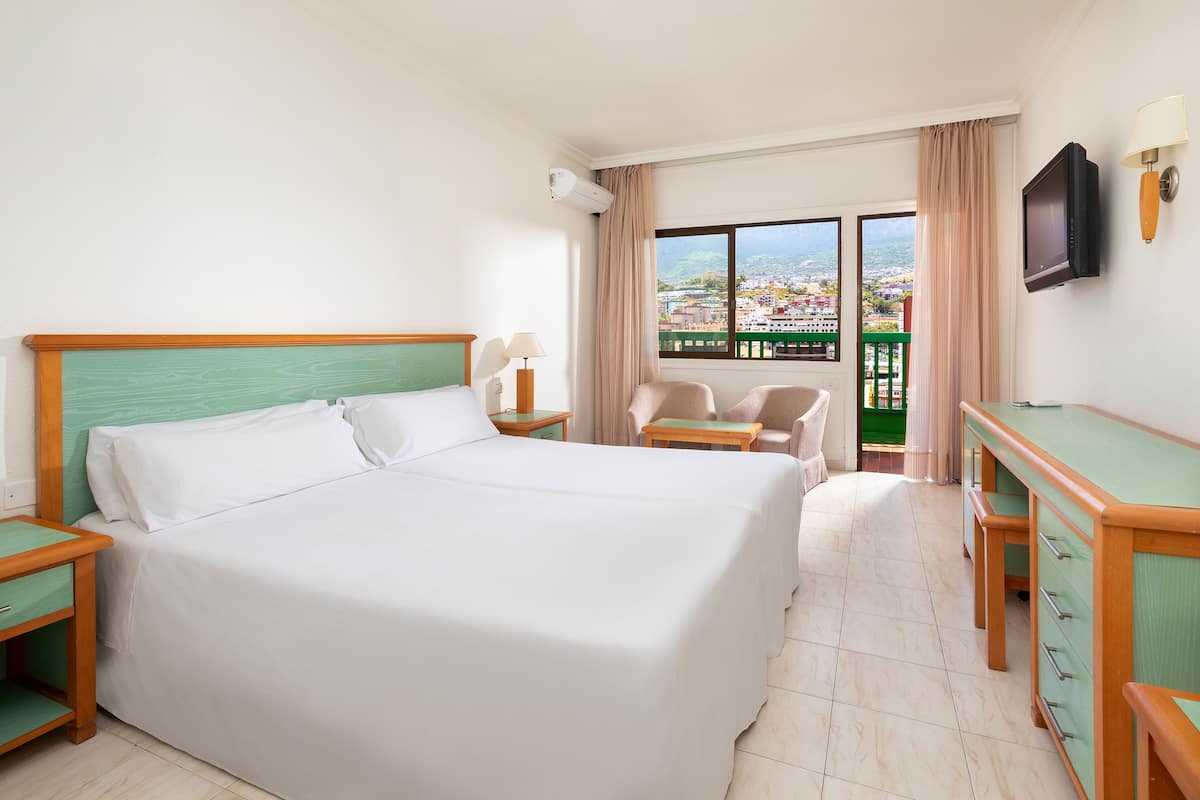 Ein Hotelzimmer mit einem großen Bett, einem Schreibtisch, einem Sessel, einem Fernseher und einem Balkon mit Blick auf die Stadt.