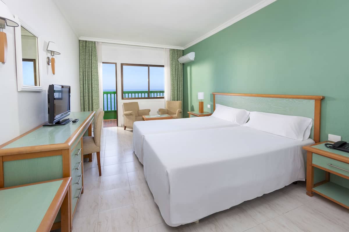 Ein Hotelzimmer mit einem großen Bett, einem Schreibtisch mit Stuhl, einem Fernseher und Blick auf das Meer.