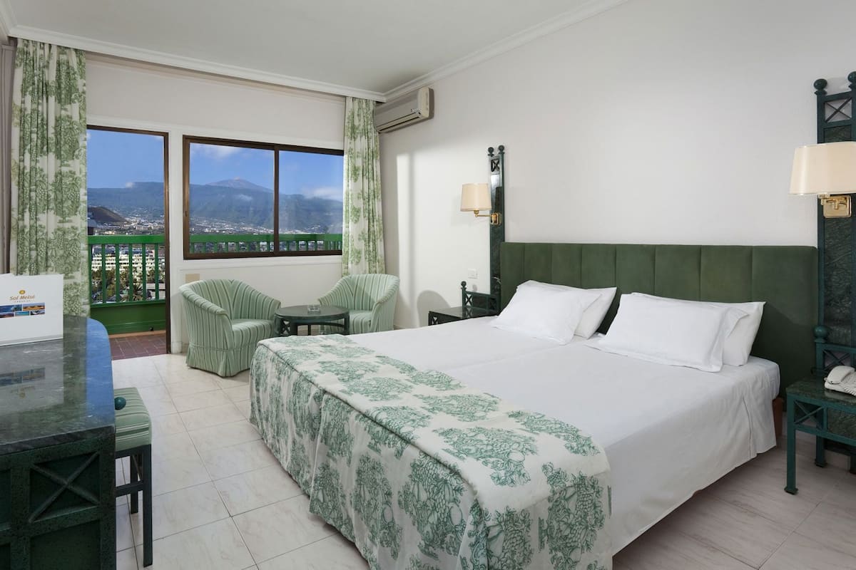 Ein Hotelzimmer mit Bett, zwei Sessel, einem kleinen Tisch, einem Schreibtisch, einem Telefon und Blick auf Stadt und Berge.