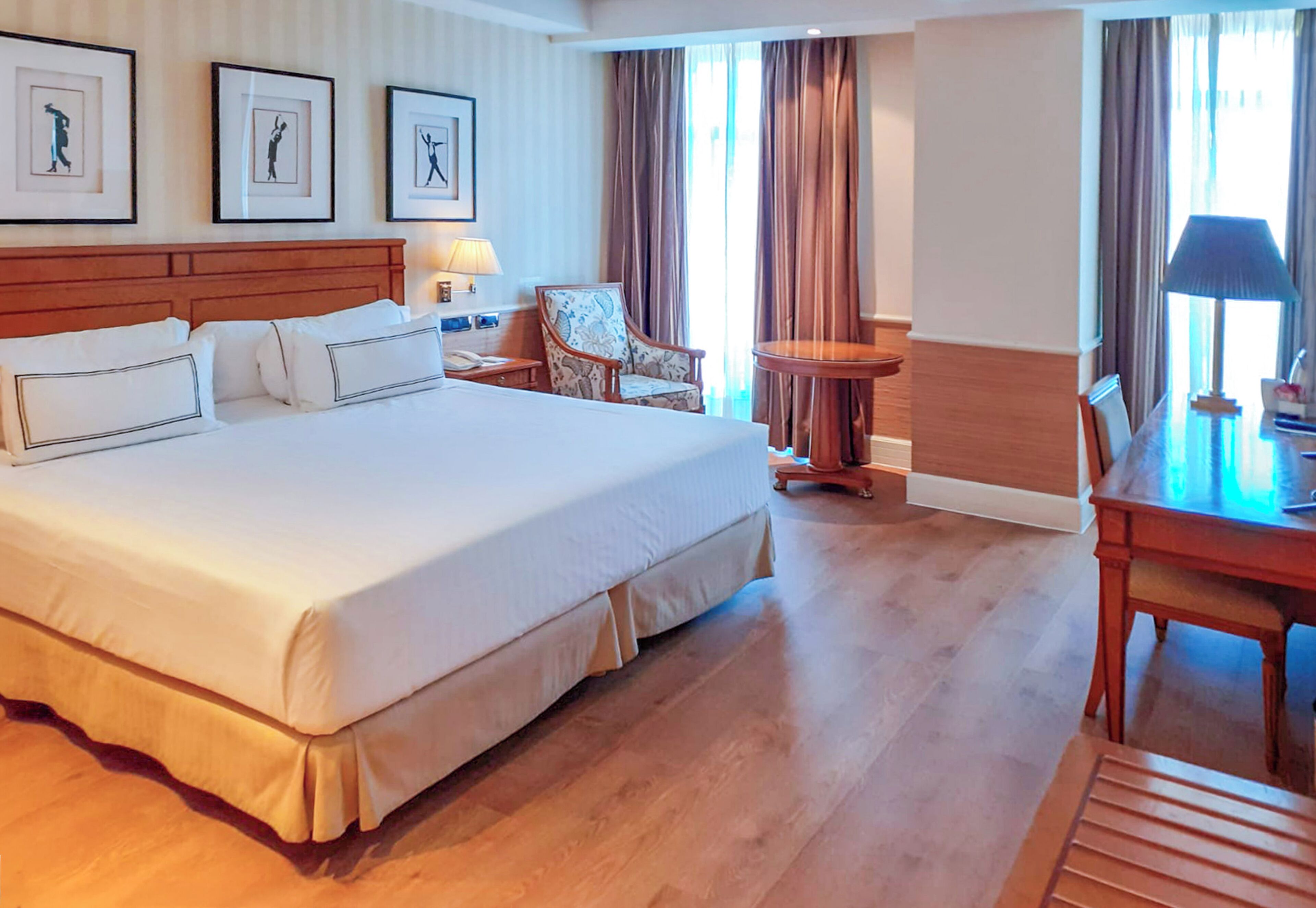 Quarto de hotel com uma cama grande, uma escrivaninha, uma cadeira e uma pequena mesa. Há cortinas e uma janela.