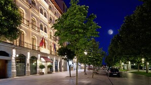 Una calle iluminada de noche con un edificio que tiene balcones y banderas, una acera bordeada de árboles y un coche a la vista.