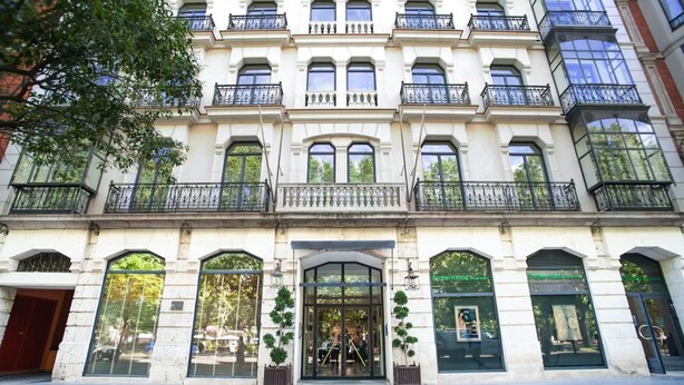 Un edificio de varios pisos con estilo arquitectónico clásico, que presenta una entrada central con puertas de vidrio, varios balcones con barandillas negras y ventanas amplias.