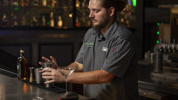 A bartender preparing a drink at a bar with shelves of bottles in the background. - Holiday Inn Wilmington by IHG (Wilmington)