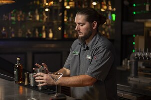 Un bartender preparando una bebida en la barra, con estantes llenos de botellas al fondo.