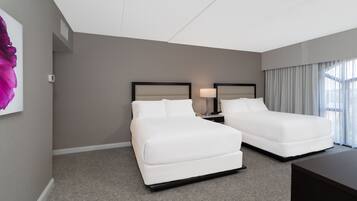 A hotel room with two beds, a nightstand with a lamp, a large window with curtains, and a painting on the wall.