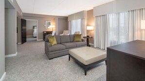 A modern hotel room with a grey sofa, a beige ottoman, a small table, and a large window with curtains. - Holiday Inn Wilmington by IHG (Wilmington)