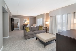 Ein modernes Hotelzimmer mit grauem Sofa, einem beigen Hocker, einem kleinen Tisch und einem großen Fenster mit Vorhängen.