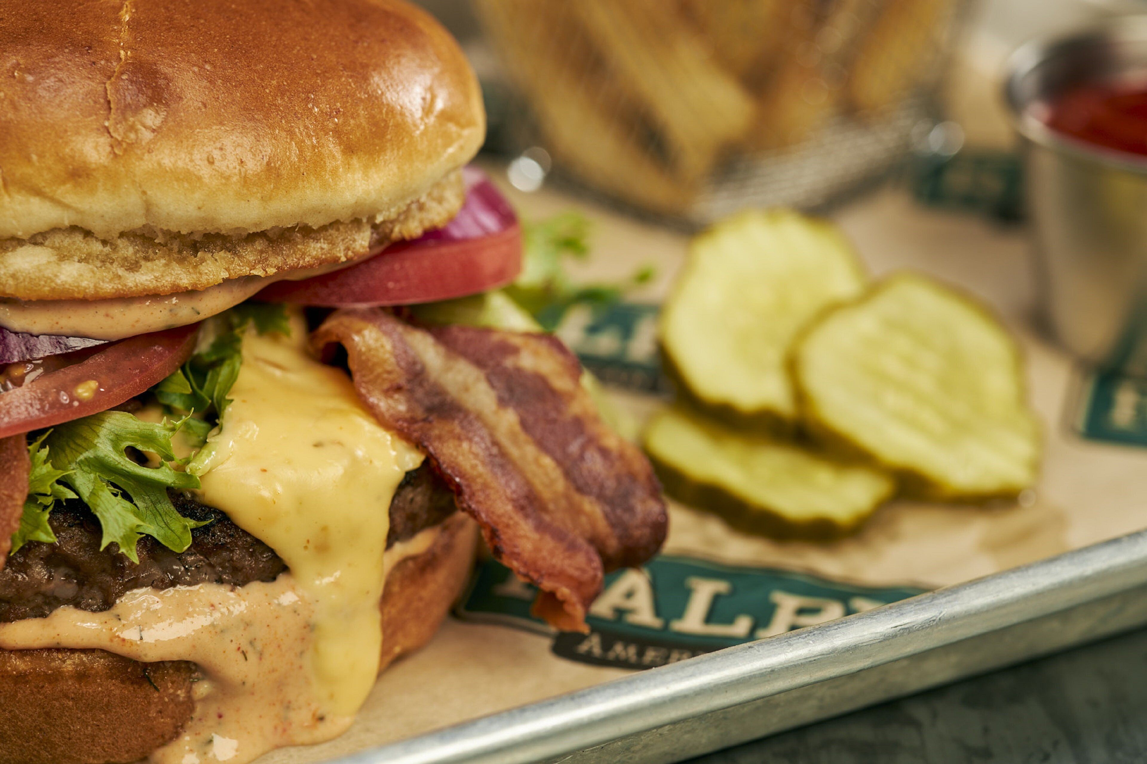 A close-up of a burger with bacon, cheese, lettuce, and tomato, served with pickle slices.