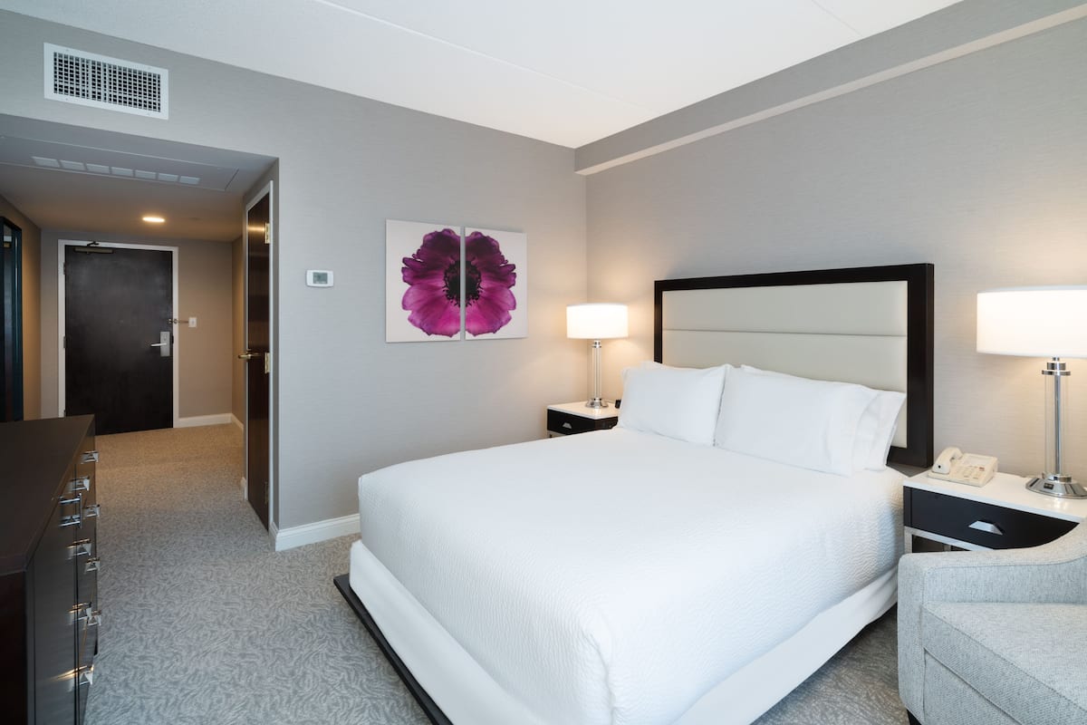 Habitación de hotel con una cama grande, dos mesitas de noche con lámparas, una obra de arte en la pared y una puerta que da a otra habitación.