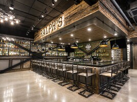 Zona de bar con un letrero con el nombre, varias luces colgantes y una variedad de botellas en estanterías.