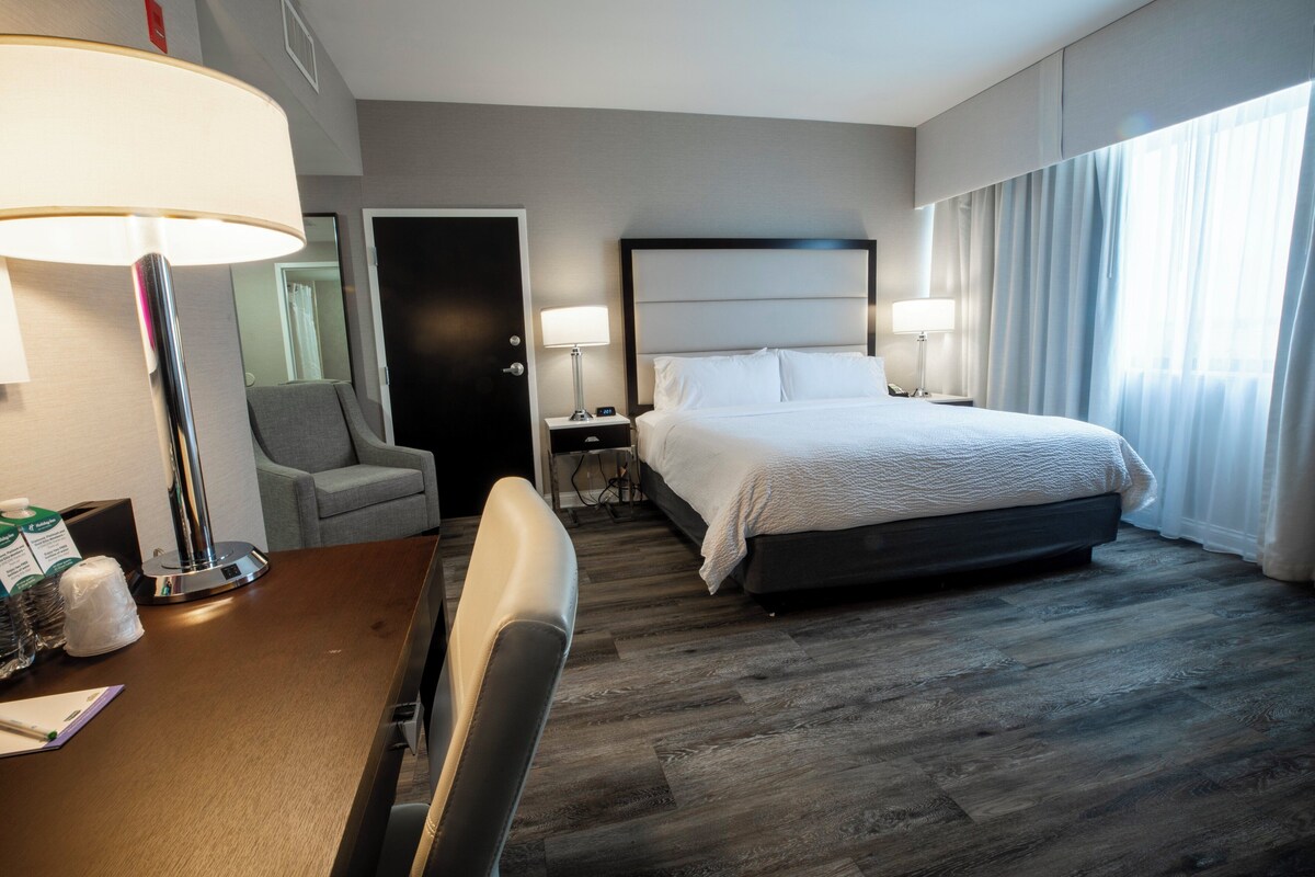 A hotel room with a bed, a desk with a chair, a lamp, and a nightstand.