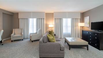 Habitación de hotel con un sofá gris, dos sillones, un televisor de pantalla plana, un mueble y un ventanal grande con cortinas.