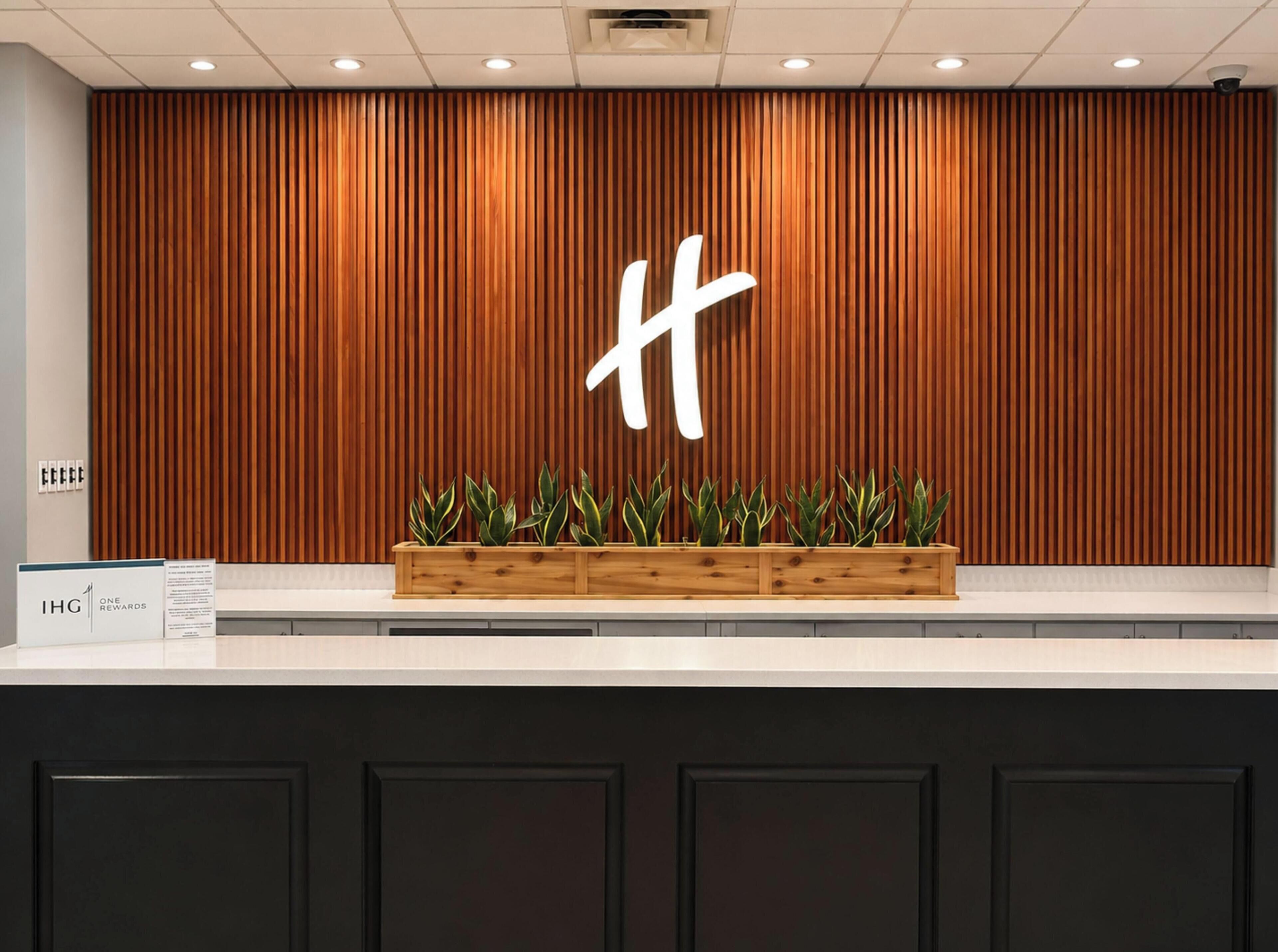 A reception desk with a wooden panel wall, a large illuminated 'H' sign, and a planter with green plants.