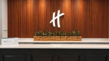 A reception desk with a wooden panel wall, a large illuminated 'H' sign, and a planter with green plants.