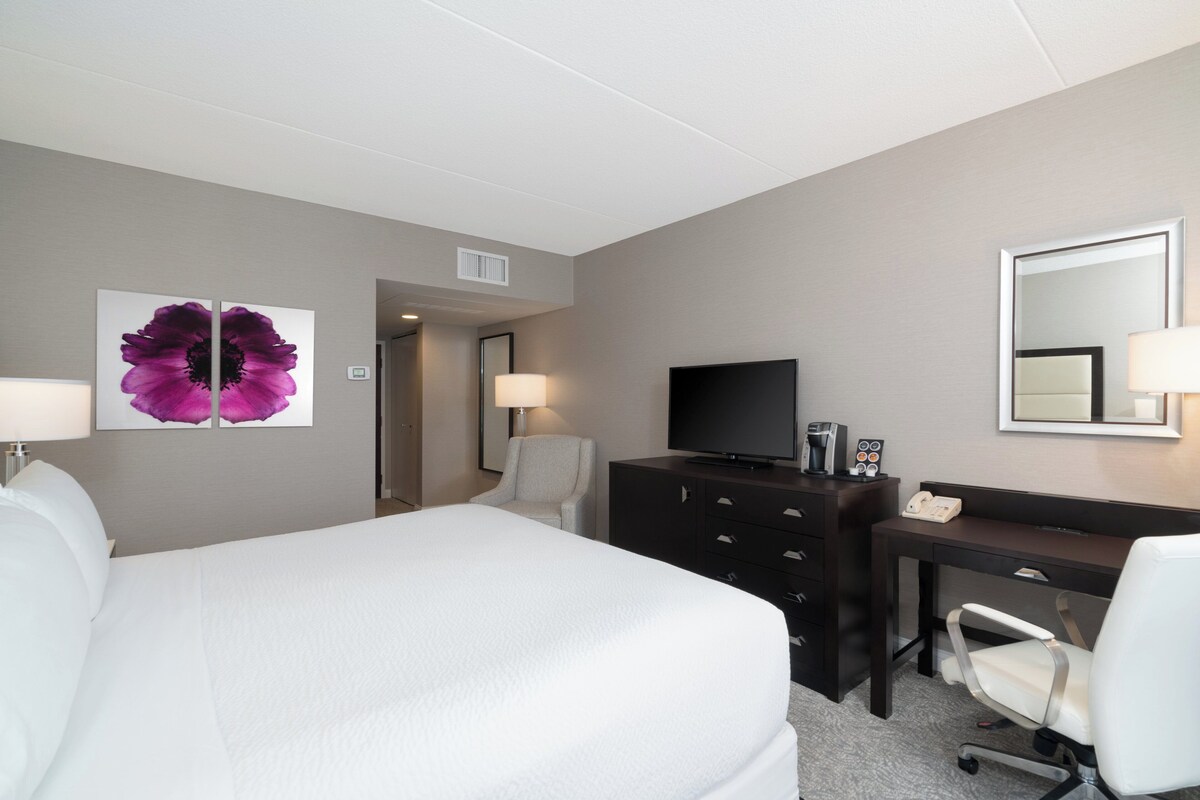 A hotel room with a large bed, a desk with a television, a chair, and a dresser.