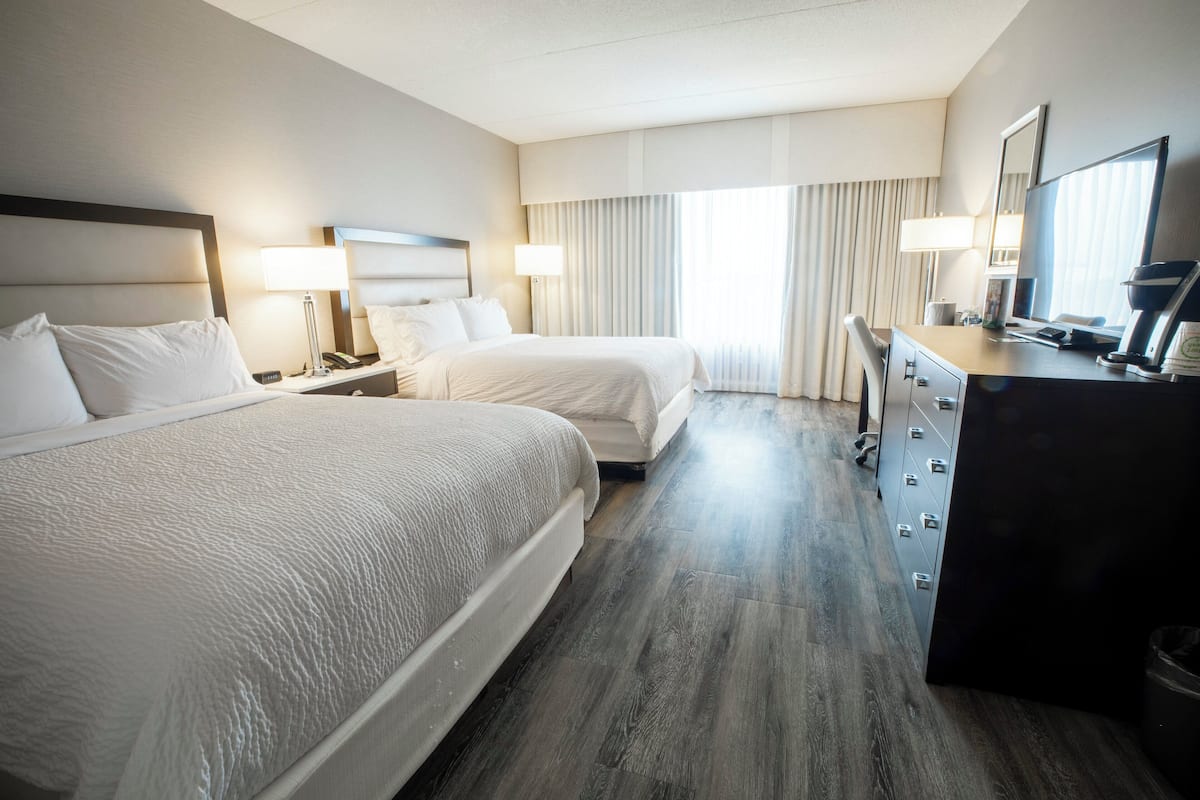 Habitación de hotel con dos camas, un escritorio, una silla, un aparador y un amplio ventanal con cortinas.