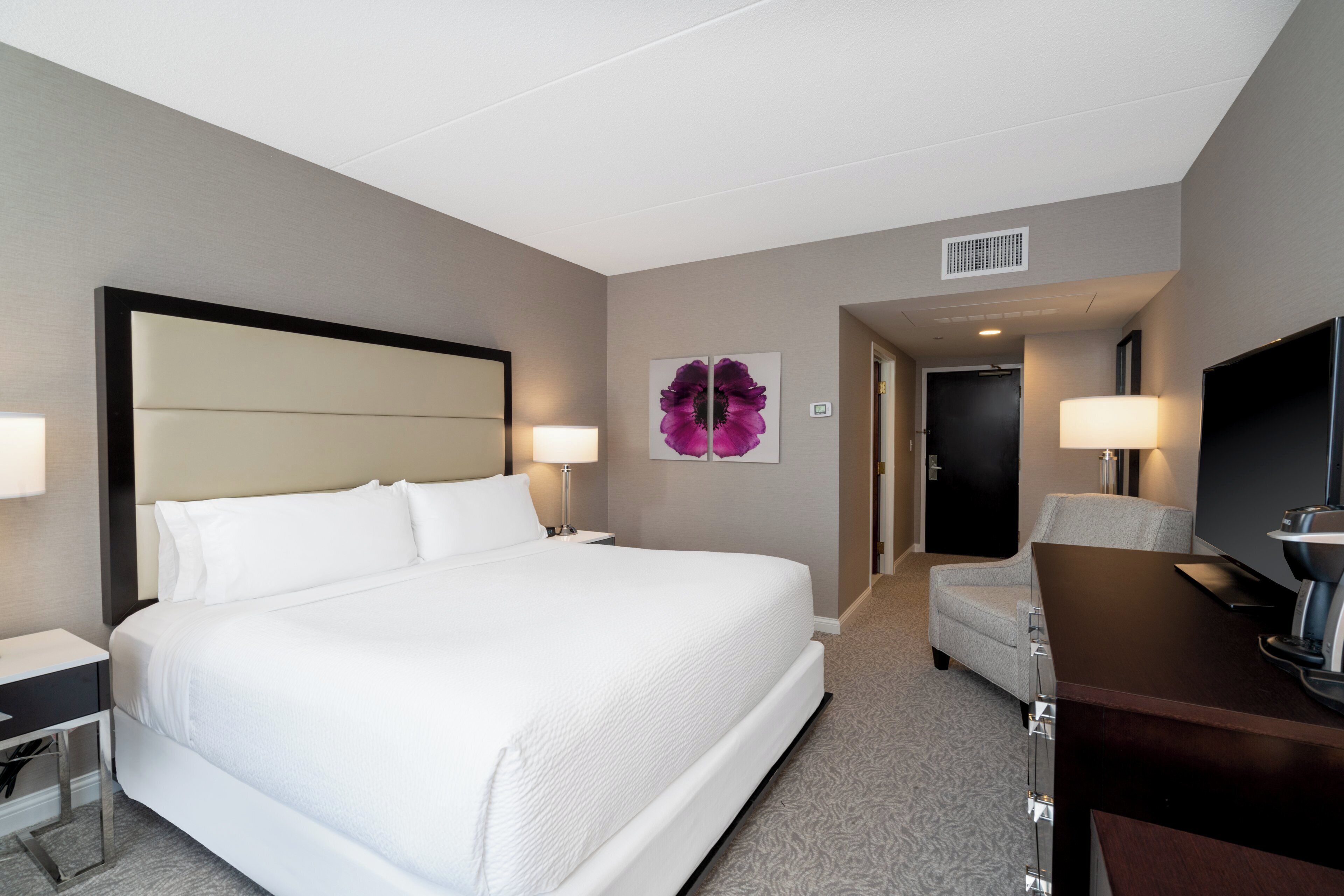A hotel room with a large bed, a desk, a chair, a TV, and a door leading to another room.