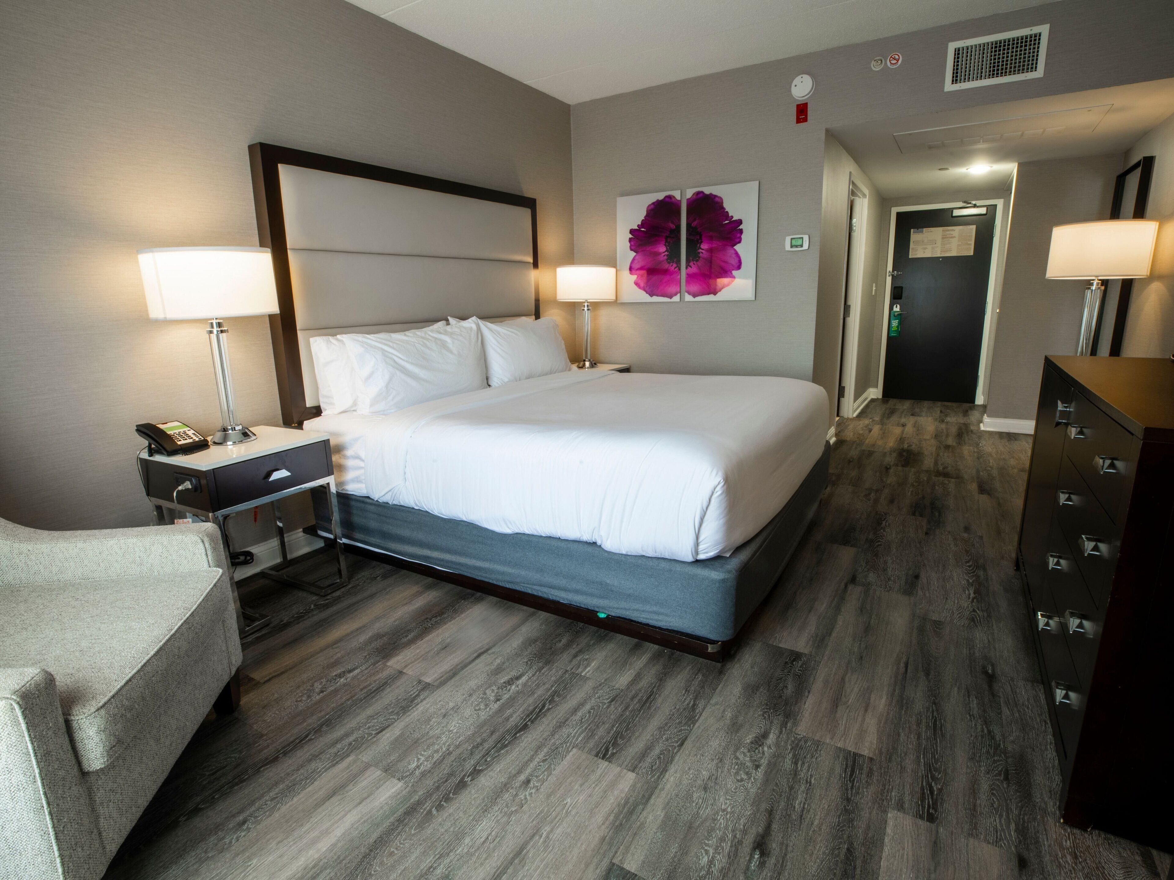 Una habitación de hotel con una cama grande, mesitas de noche con lámparas, una silla, un aparador y una puerta que da a otra habitación.