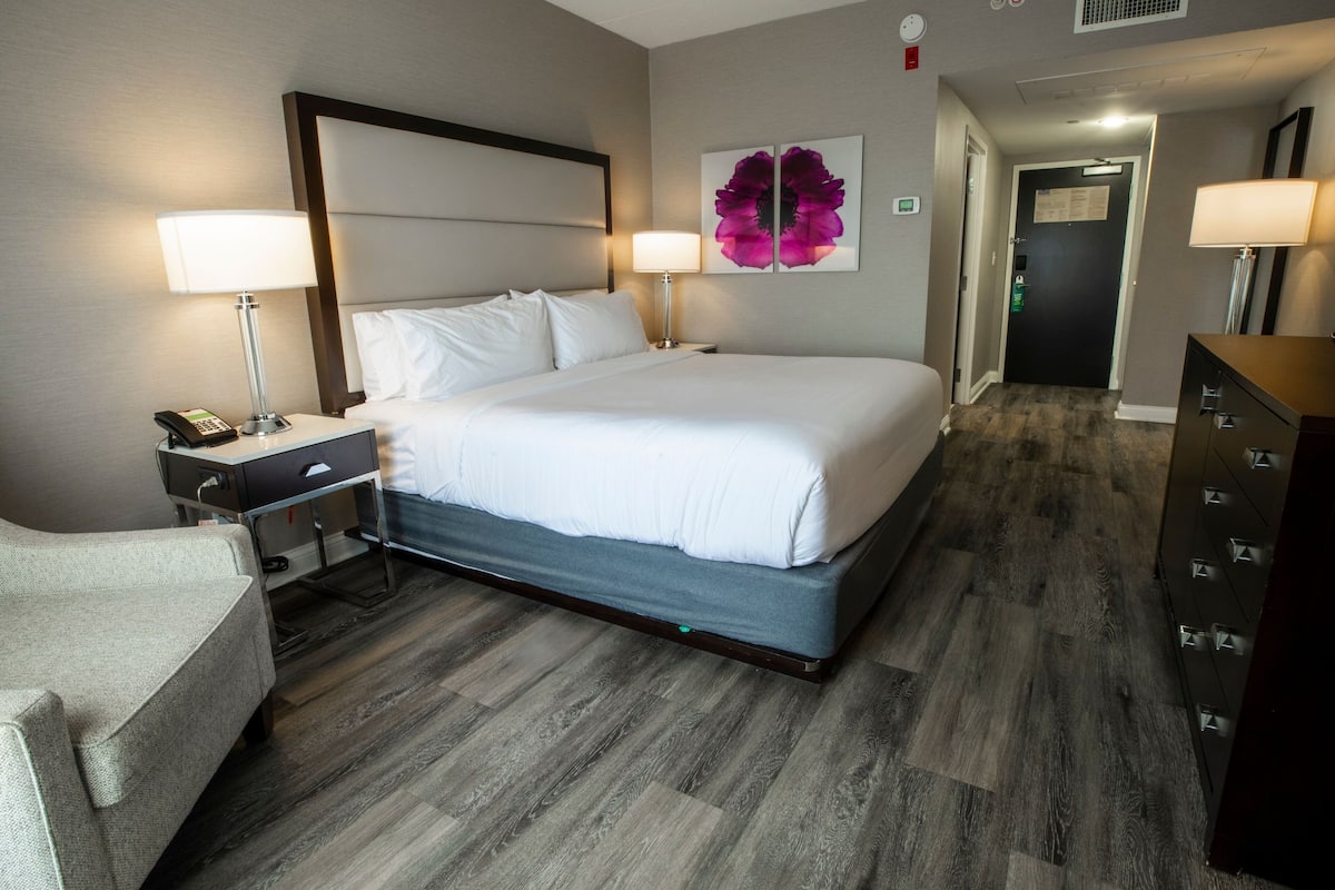 Una habitación de hotel con una cama grande, mesitas de noche con lámparas, una silla, un aparador y una puerta que da a otra habitación.