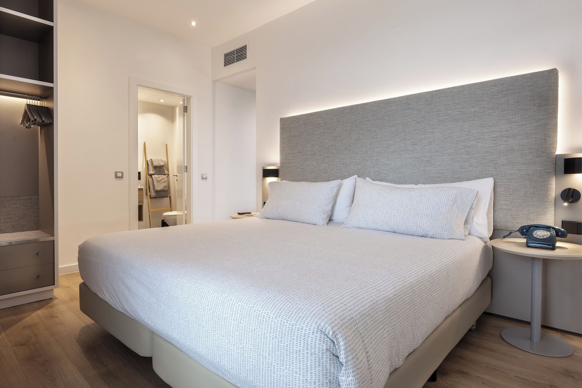 A modern hotel room with a large bed, a bedside table with a lamp, and a wardrobe.