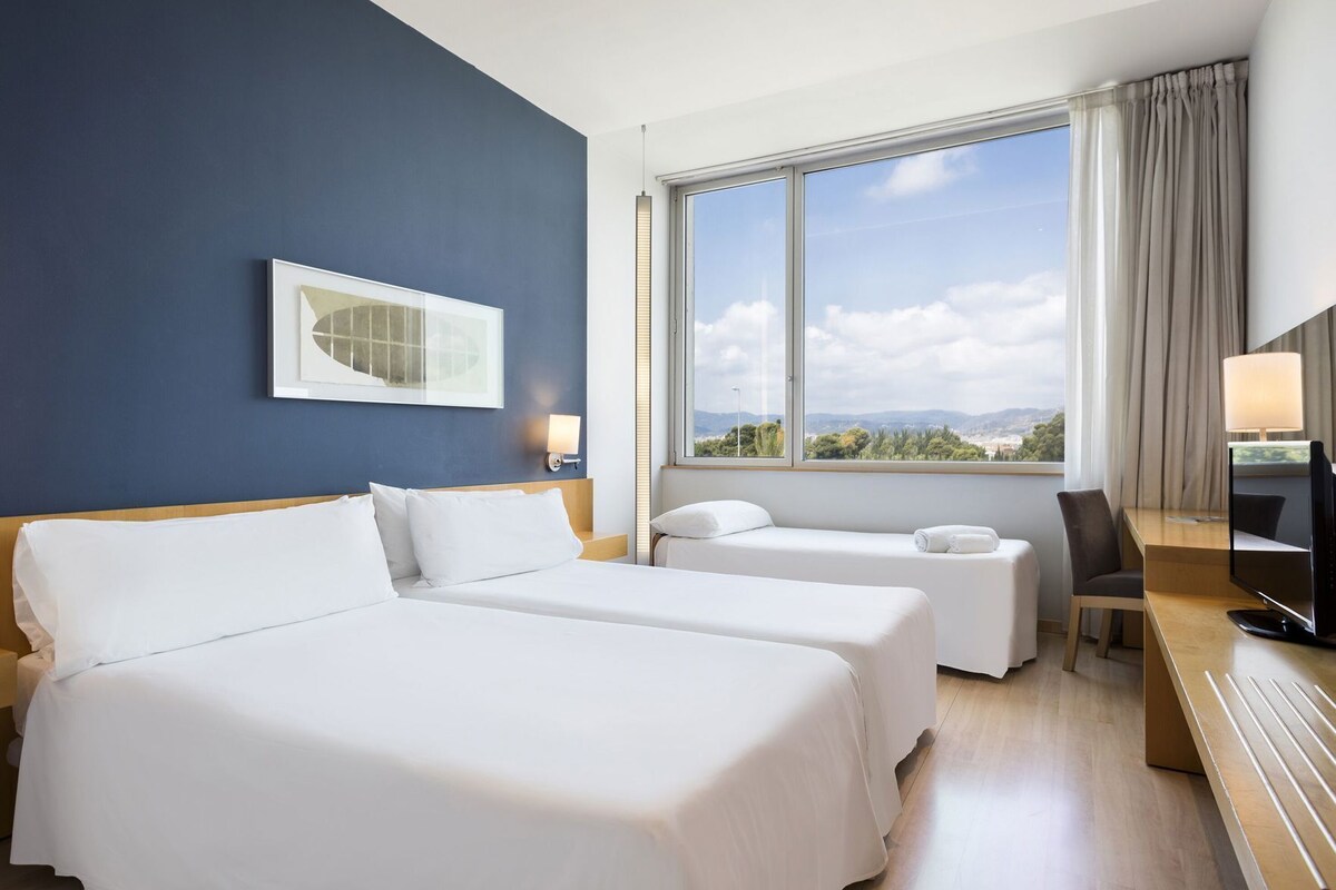 Una camera d'albergo con due letti, una scrivania, una sedia, una televisione, una finestra con vista e una lampada a parete.