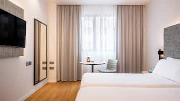 Habitación de hotel con una cama grande, una mesita pequeña, una silla, un televisor montado en la pared y una ventana con cortinas.