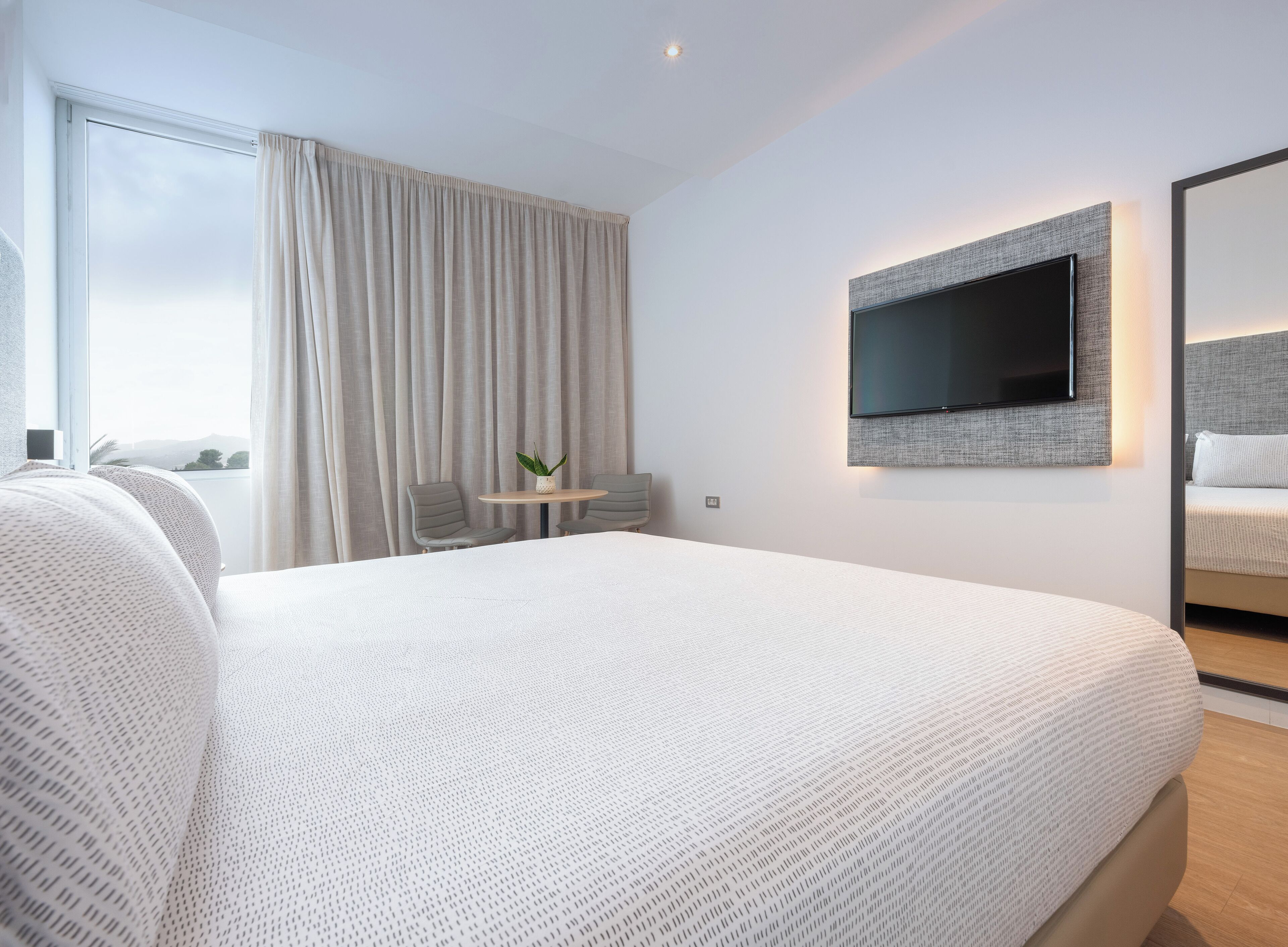 Una habitación de hotel moderna con una cama grande, un televisor de pantalla plana montado en la pared, una pequeña mesa con sillas y vistas al exterior a través de la ventana.
