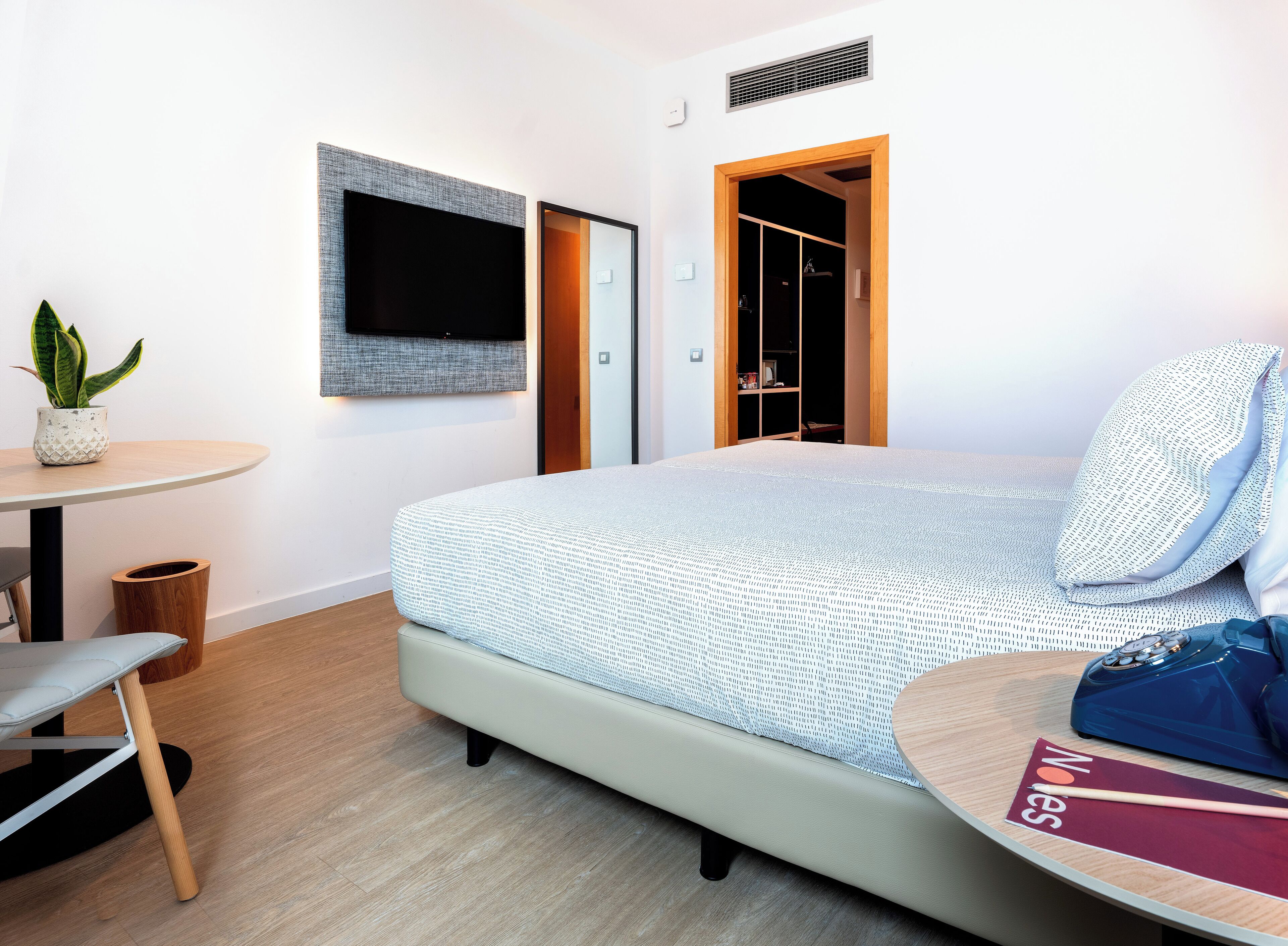 Habitación de hotel moderna con cama, una mesita con silla, una planta en maceta, un televisor montado en la pared y un armario con espejo.