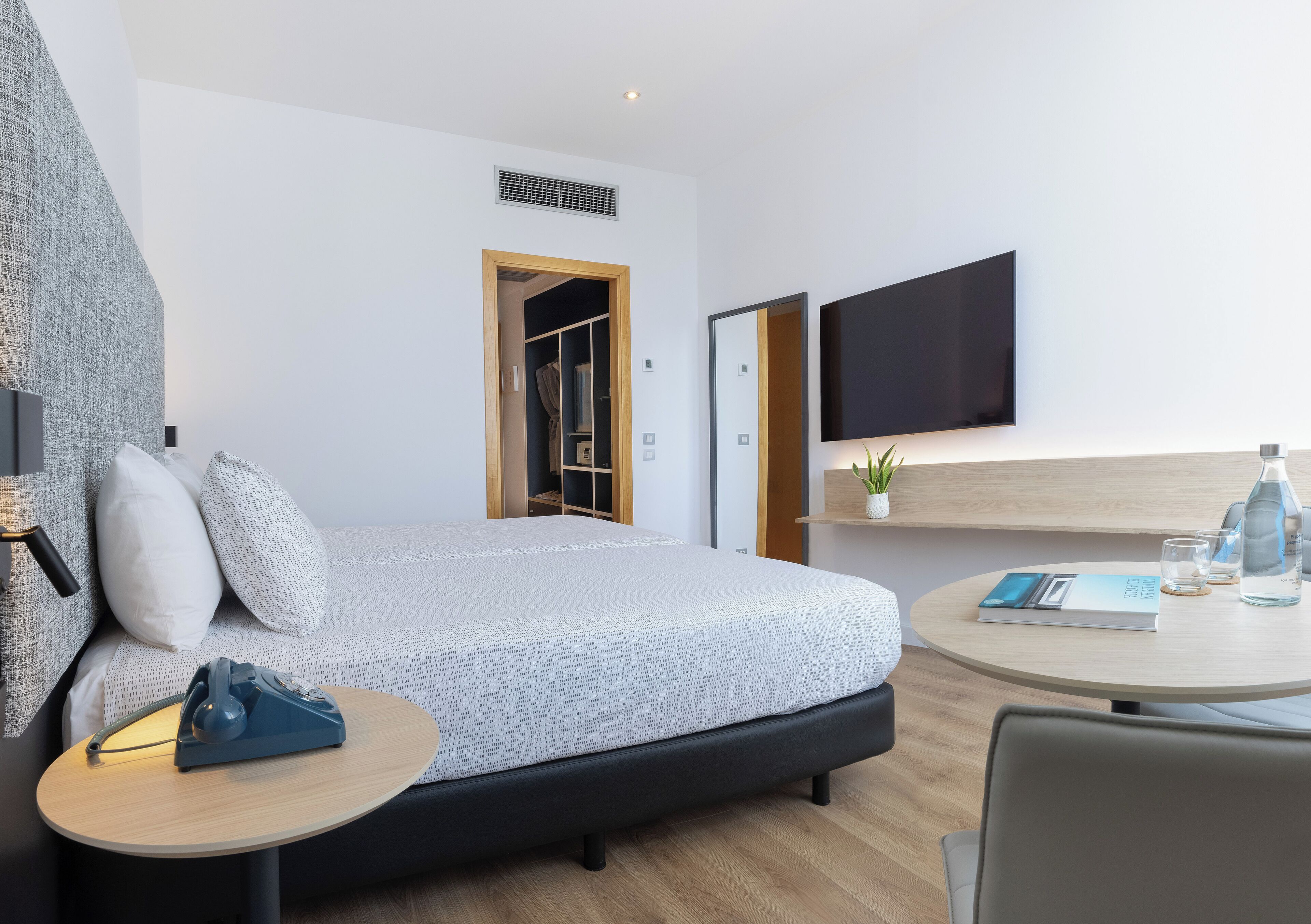 Una habitación de hotel moderna con cama, mesita de noche, silla, televisor y una pequeña mesa con un libro y vasos.